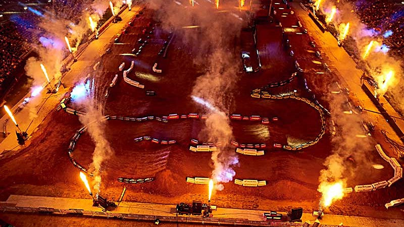 A stadium Supercross show under lights, with pyrotechnics ringing the track.
